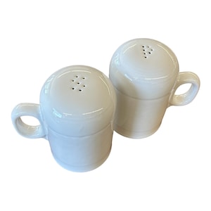Puede incluir: Un par de saleros y pimenteros de cerámica blanca. Cada salero y pimentero tiene una parte superior redondeada con múltiples agujeros y un asa pequeña. Los saleros y pimenteros están diseñados para parecerse a tazas pequeñas.