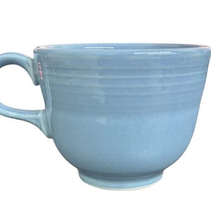Puede incluir: Una taza de cerámica de color azul claro con un acabado brillante y una serie de estrías horizontales que rodean la parte superior del cuerpo. La taza tiene un asa curva en el lado izquierdo y una base circular, con una superficie lisa y sólida.