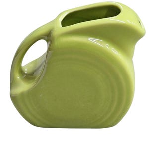 Può includere: Una brocca in ceramica verde chiaro con un manico curvo e un beccuccio. La brocca ha un corpo arrotondato e una finitura liscia e lucida. L'apertura è rettangolare. La brocca è probabilmente vintage e adatta per servire bevande.