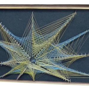 May include: A framed string art piece featuring a starburst pattern created with yellow and light blue threads stretched across small nails on a dark charcoal background, enclosed within a thin metallic gold-toned rectangular frame.