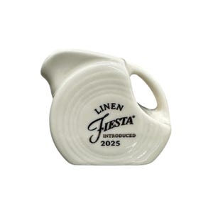 Può includere: Una brocca in ceramica bianca con manico e beccuccio curvi. La brocca presenta le scritte "Linen Fiesta" e "Introduced 2025" stampate in nero sul davanti. La brocca ha una finitura liscia e lucida.