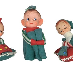 May include: Three vintage elf figurines. Each elf has a painted face and is wearing a green and red outfit. The elves are sitting on green cushions with red skirts. The outfits have white and green striped accents.