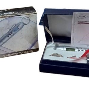 Op de afbeelding: Een zilveren digitale weegschaal in de vorm van een pen met een zwarte en zilveren doos. De weegschaal heeft een klein LCD-scherm en een haak om voorwerpen aan te hangen. De doos bevat een gebruiksaanwijzing en een klein rood lint.