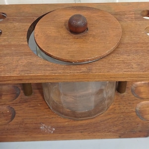 Könnte beinhalten: Holz-Pfeifenständer mit einem Glasgefäß für Tabak. Der Ständer hat acht Löcher für Pfeifen und ein Fach mit Deckel für das Gefäß. Das Gefäß ist aus klarem Glas und hat einen Holzdeckel mit einem Knopf.