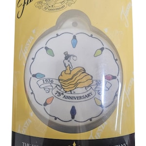 May include: A Fiesta 75th Anniversary ornament in a clear plastic package. The white ornament features a woman in a yellow dress, surrounded by colorful lights. The packaging includes the Fiesta logo and text from The Homer Laughlin China Company.