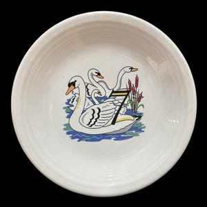 May include: White ceramic bowl with a blue and green design of four white swans swimming in a pond with red reeds. The swans are stylized and have black markings. The bowl has a white rim.