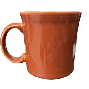 Puede incluir: Una taza de cerámica rojo-naranja brillante con un asa curva. La taza tiene un borde ligeramente ensanchado y una superficie lisa y sin adornos. La taza está diseñada para bebidas calientes.