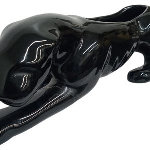 May include: A glossy black ceramic panther figurine in a crawling pose. The panther has a sleek, curved body with a hollowed-out back, possibly for holding items. The figurine is a decorative item.