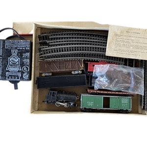 Puede incluir: Un set de tren modelo vintage en su caja original. El set incluye una locomotora negra, tres vagones de carga, un transformador y piezas de vía. La caja es marrón con texto blanco que dice "Mini-Gauge Railroad Hobby Transformer".