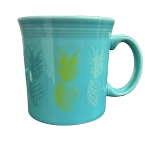 May include: A turquoise ceramic mug with a curved handle. The mug features three pineapple designs in shades of green and white. The mug has a smooth, glossy finish and is suitable for hot or cold beverages.