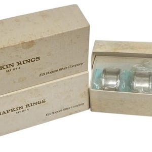 May include: A set of four silver napkin rings in a beige box. The box is labelled "NAPKIN RINGS SET OF 4". The rings are cylindrical and shiny. The box is open, showing the rings inside, which are packaged in clear plastic.