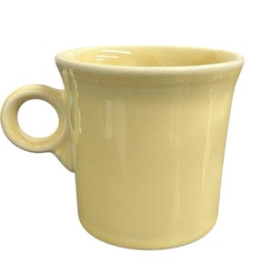 Puede incluir: Una taza de cerámica amarillo pálido con un asa redondeada. La taza tiene un borde ligeramente ensanchado y un acabado liso y brillante. La taza está diseñada para bebidas calientes.