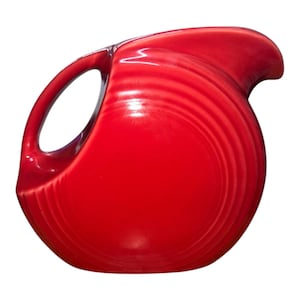 May include: A vibrant red ceramic pitcher with a rounded body and a distinctive handle. The pitcher features a curved spout and a series of concentric ridges that encircle the body. The handle is a large, open loop. The pitcher is a solid, glossy red.