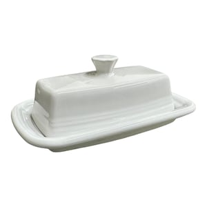 May include: A white ceramic butter dish with a lid and a small handle. The dish is rectangular with rounded edges and a slightly raised base. The lid has a small, conical handle. The overall design is simple and elegant.