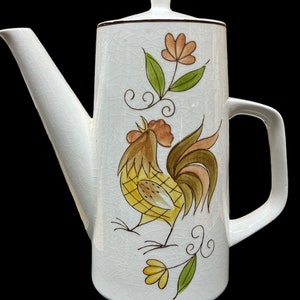 Könnte beinhalten: Weiße Kaffeekanne aus Keramik mit Hahn und Blumenmuster. Der Hahn ist gelb, braun und grün. Die Blumen sind orange und grün.