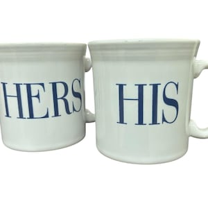 May include: Two white ceramic mugs with blue text. One mug reads "HERS" and the other reads "HIS". Both mugs have a handle and a simple design. The mugs are likely for beverages.