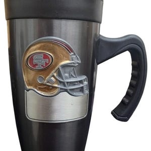 May include: A stainless steel travel mug with a black handle and lid. The mug features a gold and red football helmet emblem. The mug is designed to keep beverages hot or cold.