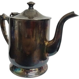 May include: A silver-colored metal coffee pot with a lid, handle, and spout. The pot has a cylindrical body with a rounded base and a curved spout. The lid is flat with a small knob on top. The handle is attached to the side of the pot.