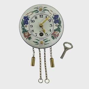 Puede incluir: Una esfera de reloj vintage con fondo blanco, números negros y diseños florales en azul, rojo y verde. El reloj tiene manecillas doradas, una llave y cadenas colgantes con pesas. La palabra "GERMANY" está impresa en la parte inferior.
