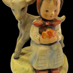 May include: A porcelain figurine of a girl in a red and white dress holding two small birds. She is sitting next to a white goat with brown eyes.