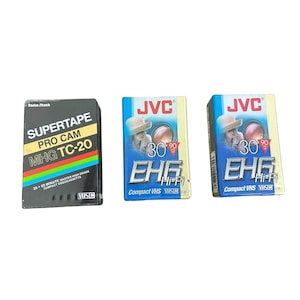Könnte beinhalten: Drei Vintage-VHS-Videokassetten. Eine ist schwarz mit dem Text "SUPERTAPE PRO CAM MHG TC-20". Die anderen beiden sind blau mit dem Text "JVC" und "EHG Hi-Fi Compact VHS". Jede Kassette hat ein anderes Design.