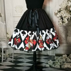May include: A black skirt featuring a wide hem band printed with a white diamond pattern, black card suit symbols, and colorful illustrations of characters including a queen and a rabbit. The skirt has a solid black waistband with a tied ribbon bow.