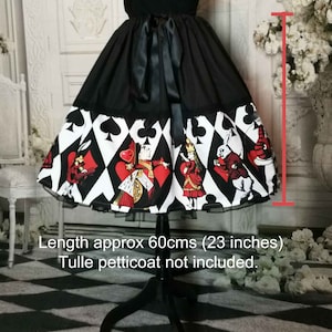 May include: A black skirt featuring a wide hemline print of playing card suits and Alice in Wonderland characters. The skirt has a black satin ribbon tie at the waist and measures 23 inches in length. The garment is displayed on a mannequin.