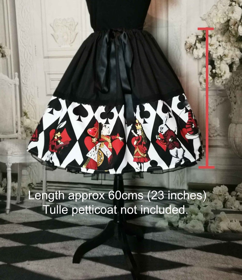May include: A black skirt featuring a wide hemline print of playing card suits and Alice in Wonderland characters. The skirt has a black satin ribbon tie at the waist and measures 23 inches in length. The garment is displayed on a mannequin.