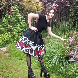 May include: A black short-sleeved top and a flared skirt featuring a playing card suit pattern in red, black, and white. The outfit includes a matching top hat with a red ribbon, black vertical striped tights, and black platform high-heeled shoes.