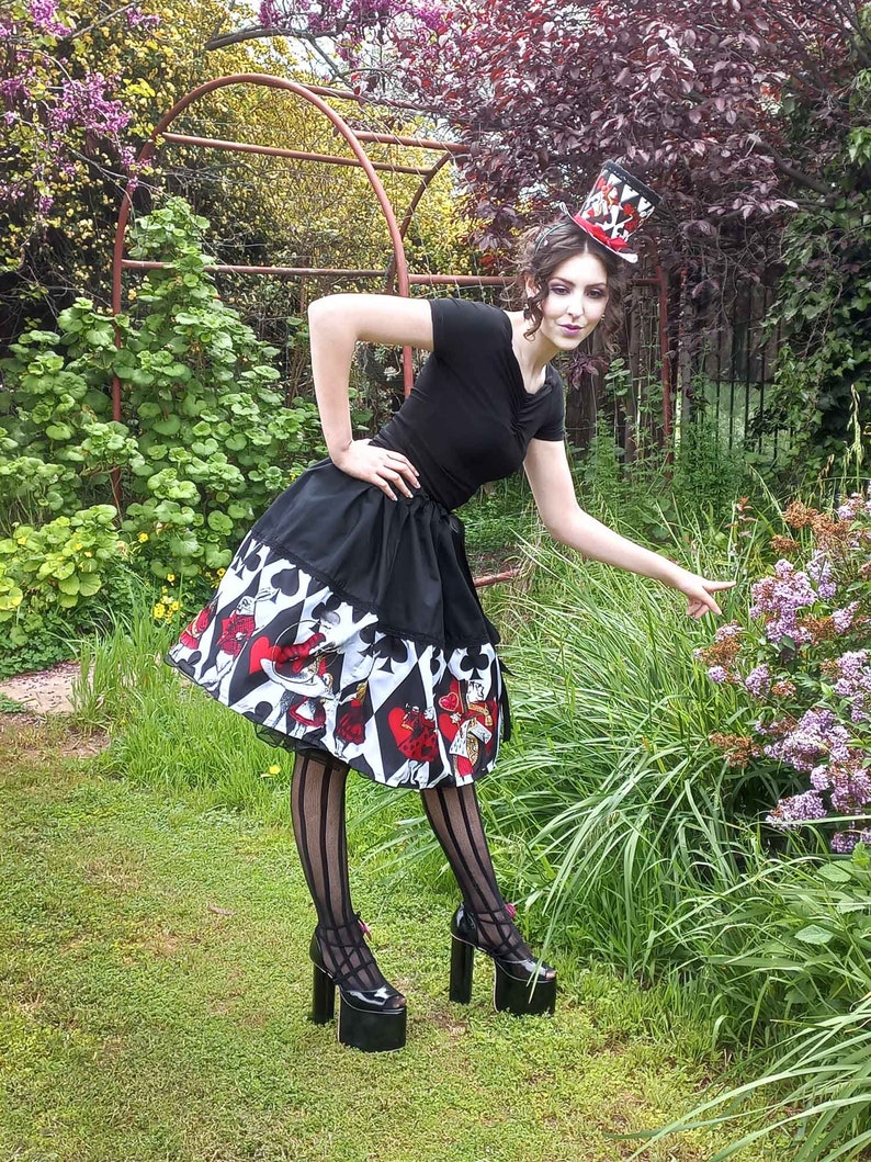 May include: A black short-sleeved top and a flared skirt featuring a playing card suit pattern in red, black, and white. The outfit includes a matching top hat with a red ribbon, black vertical striped tights, and black platform high-heeled shoes.