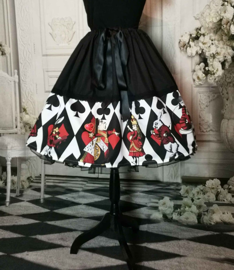 May include: A black skirt featuring a wide hem band printed with a white diamond pattern, black card suit symbols, and colorful illustrations of characters including a queen and a rabbit. The skirt has a solid black waistband with a tied ribbon bow.