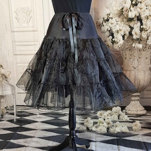 May include: A black knee-length skirt featuring a smooth waistband with a tied ribbon bow at the center front. The skirt is constructed with multiple tiers of sheer, ruffled tulle fabric that create a voluminous, layered silhouette.