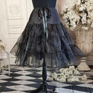 May include: A black knee-length skirt featuring a smooth waistband with a tied ribbon bow at the center front. The skirt is constructed with multiple tiers of sheer, ruffled tulle fabric that create a voluminous, layered silhouette.