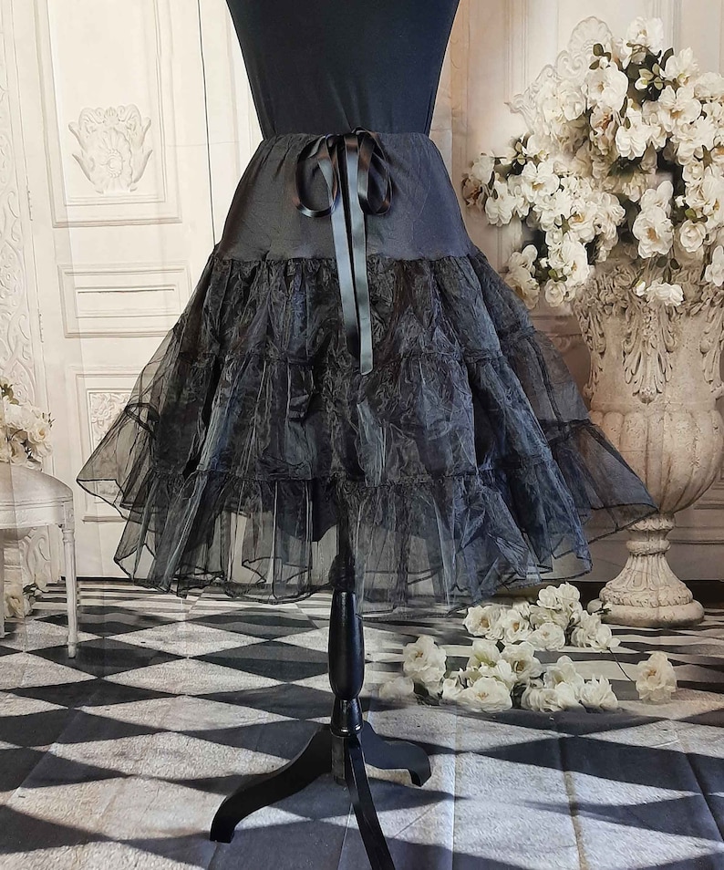 May include: A black knee-length skirt featuring a smooth waistband with a tied ribbon bow at the center front. The skirt is constructed with multiple tiers of sheer, ruffled tulle fabric that create a voluminous, layered silhouette.