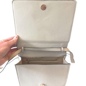 K&ouml;nnte beinhalten: Ge&ouml;ffnete, cremefarbene Handtasche mit rechteckiger Form. Die Tasche hat einen Klappverschluss mit goldfarbenem Verschluss und einen langen, d&uuml;nnen Riemen. Der Innenraum ist in einem helleren Cremeton gehalten.