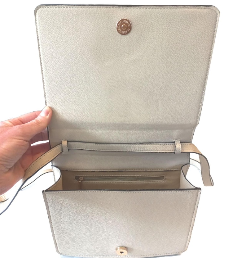 K&ouml;nnte beinhalten: Ge&ouml;ffnete, cremefarbene Handtasche mit rechteckiger Form. Die Tasche hat einen Klappverschluss mit goldfarbenem Verschluss und einen langen, d&uuml;nnen Riemen. Der Innenraum ist in einem helleren Cremeton gehalten.
