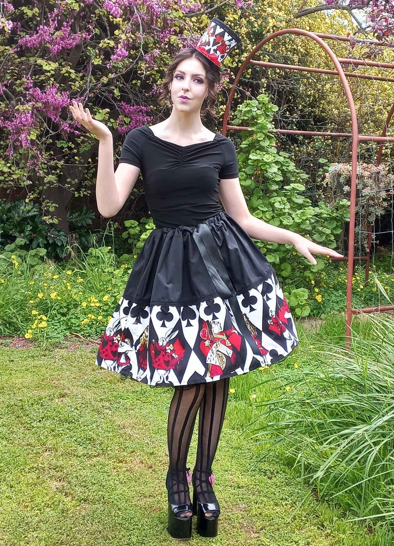 May include: A black short-sleeved top paired with a flared skirt featuring a white, black, and red playing card suit pattern. The outfit includes a matching miniature top hat adorned with card motifs, and black vertical striped tights with platform shoes.