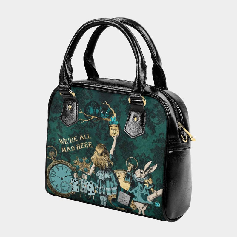 May include: Black leather handbag with a green and gold print featuring Alice in Wonderland characters, including the Cheshire Cat, the Mad Hatter, and the White Rabbit. The text "We're All Mad Here" is printed in gold on the bag.