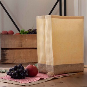 May include: Two beige canvas tote bags with brown burlap trim. The bags are sitting on a wooden table with a red and white checkered cloth and grapes and a peach on top of the cloth.