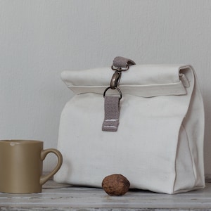 Op de afbeelding: Een witte canvas lunchtas met een bruine band en een metalen sluiting. De tas is bovenaan gevouwen en heeft een bruine stoffen handvat. Een bruine keramische mok en een walnoot bevinden zich op de voorgrond.