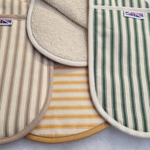 Può includere: Primo piano di diversi guanti da forno con motivi a righe in vari colori. Uno è beige con strisce marroni, un altro è giallo con strisce bianche e un terzo è verde con strisce bianche. Ogni guanto ha una piccola etichetta con la Union Jack.