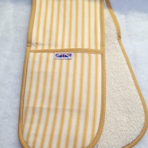 Può includere: Un doppio guanto da forno a righe verticali gialle e bianche. Il guanto ha un bordo giallo e una piccola etichetta con una bandiera Union Jack. L'interno è in morbida spugna beige.
