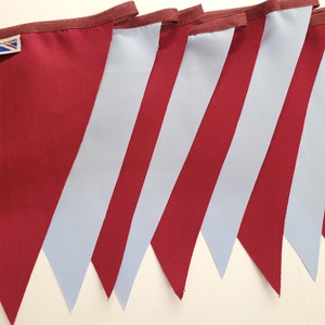 May include: A set of seven pennant flags with alternating burgundy and light blue stripes. The flags are sewn together to create a bunting. The flags have a small label with the text "Tracy Ann Designs" and a Union Jack.