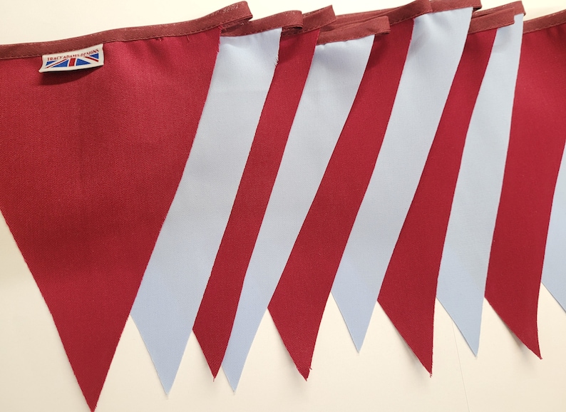 May include: A set of seven pennant flags with alternating burgundy and light blue stripes. The flags are sewn together to create a bunting. The flags have a small label with the text "Tracy Ann Designs" and a Union Jack.