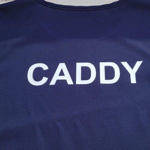 Puede incluir: Un peto azul marino con la palabra "CADDY" impresa en letras mayúsculas blancas grandes. La parte superior sin mangas tiene un escote redondo y está hecha de una tela lisa. El texto está centrado.