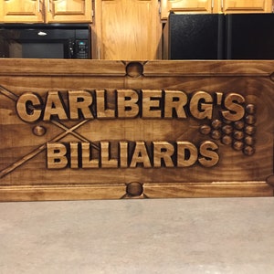 May include: A wooden sign with the text "Carlberg's Billiards" carved into it. The sign is brown and has a distressed look. There are two crossed pool cues and a cluster of pool balls carved into the sign.