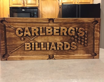 May include: A wooden sign with the text "Carlberg's Billiards" carved into it. The sign is brown and has a distressed look. There are two crossed pool cues and a cluster of pool balls carved into the sign.