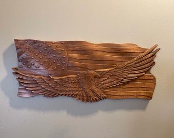 May include: A wooden carving of an American flag with an eagle flying over it. The flag is carved in a realistic style, with the stars and stripes clearly visible. The eagle is also carved in detail, with its wings spread wide.