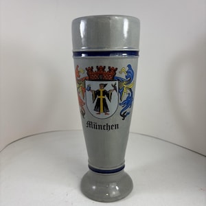 May include: A tall, tapered gray ceramic beer glass featuring a blue horizontal band near the top and bottom. The center displays a colorful coat of arms depicting a figure in a black robe with the word München printed in black gothic lettering below.