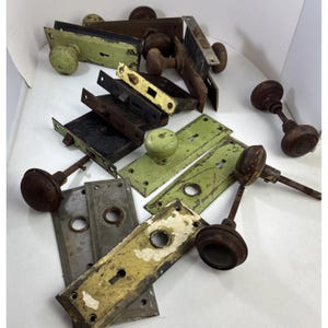 May include: A collection of antique door hardware including various metal door knobs, rectangular backplates, and mortise lock mechanisms. The items feature weathered finishes in shades of green, yellow, brown, and grey with visible signs of wear.